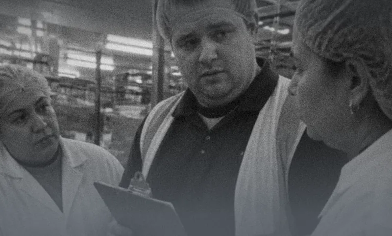 A manager with a clipboard talking to two food-factory workers in hairnets